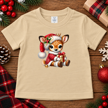 A latte colored short-sleeve kids' t-shirt featuring a cute cartoon deer character in a red Santa hat and coat holding a gift box, displayed on a rustic wooden background with Christmas decorations.