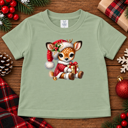 A light sage green kids' t-shirt featuring a cute cartoon Christmas deer graphic, displayed in a flat-lay arrangement with holiday decorations like pine branches, ornaments, and a plaid scarf.