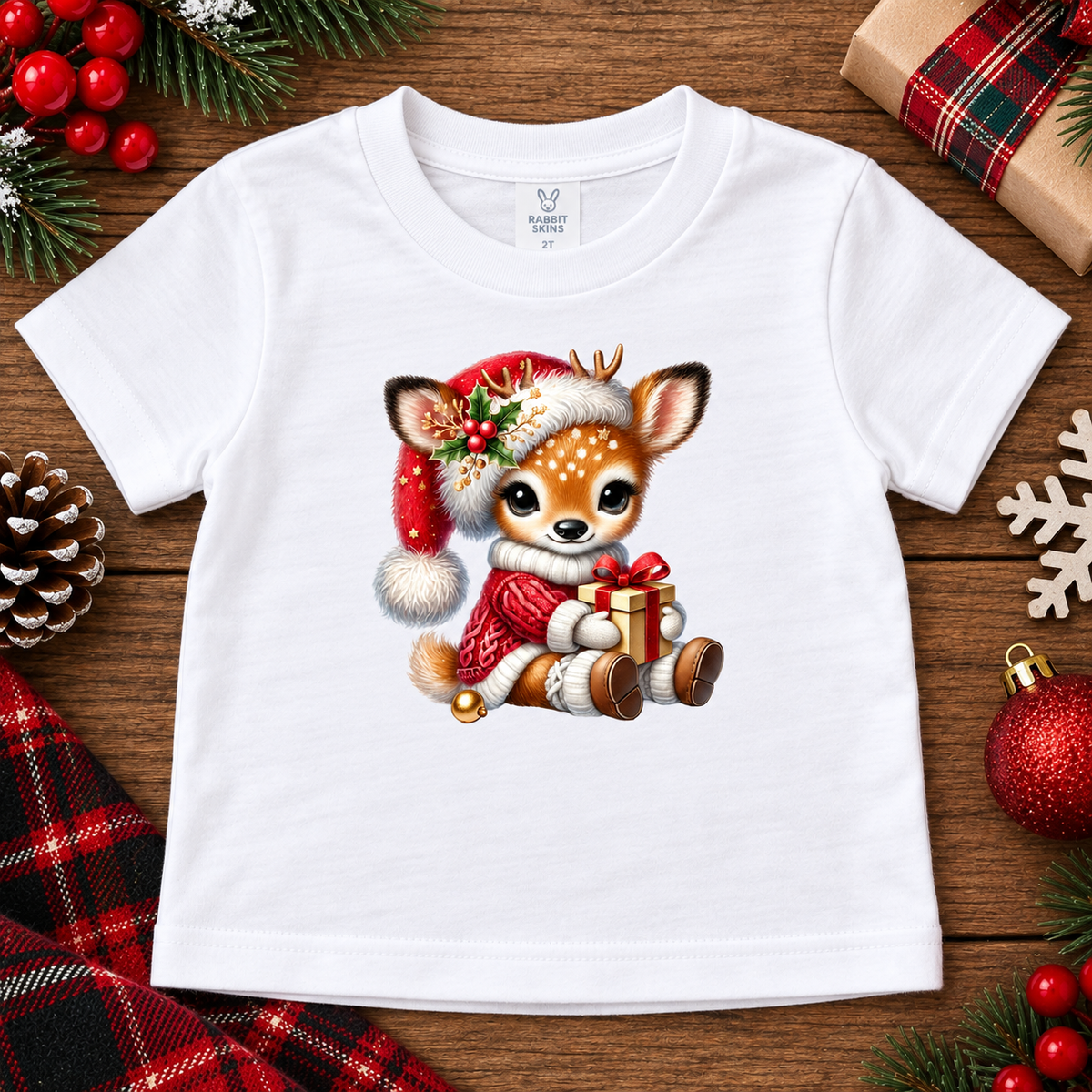 A white short-sleeve baby t-shirt with a cute cartoon deer graphic wearing a Santa hat and holding a gift, displayed on a rustic wooden background with Christmas decorations.