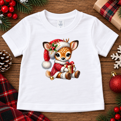 A white short-sleeve baby t-shirt with a cute cartoon deer graphic wearing a Santa hat and holding a gift, displayed on a rustic wooden background with Christmas decorations.
