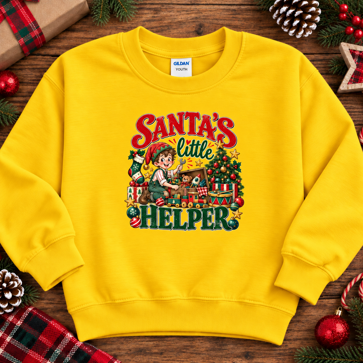 A daisy yellow crewneck sweatshirt featuring a colorful graphic print of a child elf working on a Christmas tree with the text 'Santa's Little Helper'. The sweatshirt is displayed in a flat-lay arrangement on a rustic wooden surface, surrounded by holiday decorations like pine cones, a plaid gift box, and candy canes.
