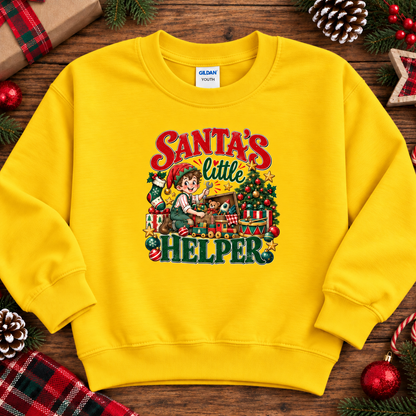 A daisy yellow crewneck sweatshirt featuring a colorful graphic print of a child elf working on a Christmas tree with the text 'Santa's Little Helper'. The sweatshirt is displayed in a flat-lay arrangement on a rustic wooden surface, surrounded by holiday decorations like pine cones, a plaid gift box, and candy canes.