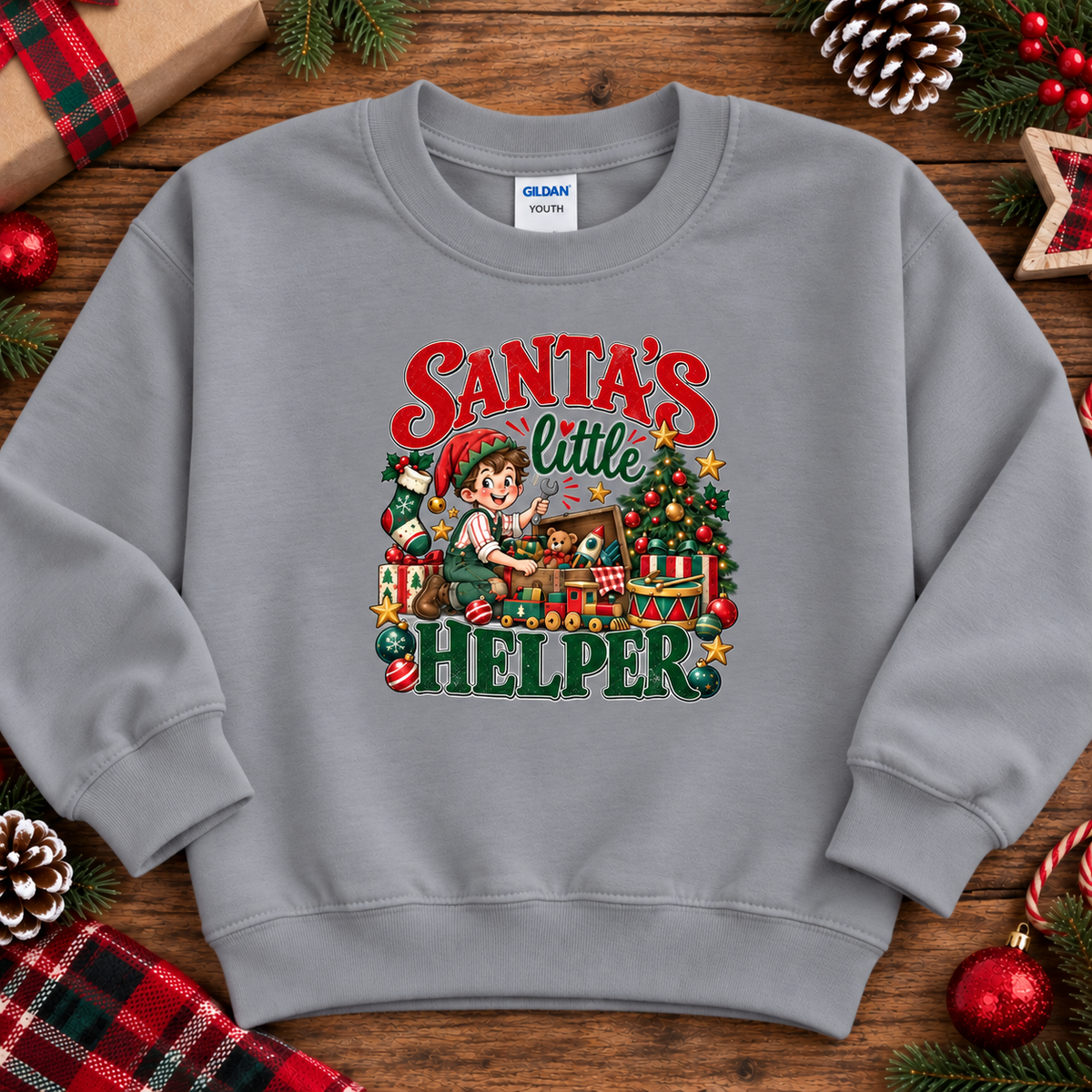 A light gray crewneck sweatshirt featuring a colorful graphic of a Christmas elf packing presents under the text 'Santa's Little Helper'. The sweatshirt is displayed in a flat-lay arrangement on a rustic wooden surface, surrounded by holiday decorations including pinecones, a wrapped gift, and red ornaments.