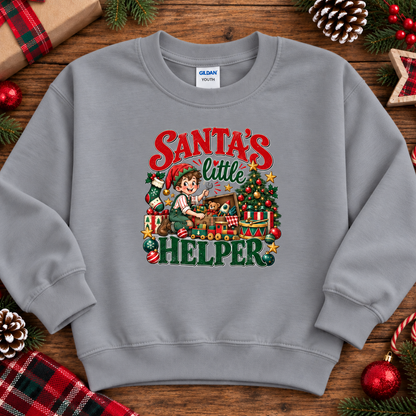 A light gray crewneck sweatshirt featuring a colorful graphic of a Christmas elf packing presents under the text 'Santa's Little Helper'. The sweatshirt is displayed in a flat-lay arrangement on a rustic wooden surface, surrounded by holiday decorations including pinecones, a wrapped gift, and red ornaments.
