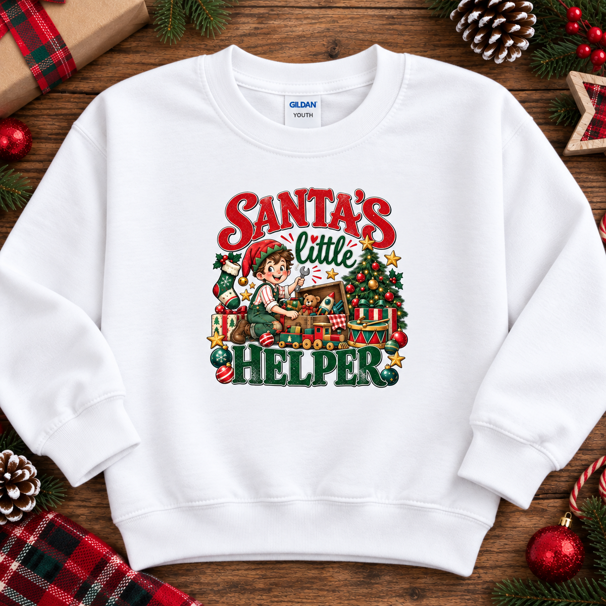 A white crewneck sweatshirt with a colorful graphic print reading 'Santa's Little Helper' featuring an elf and Christmas tree illustration, laid flat on a rustic wooden background with holiday decorations.