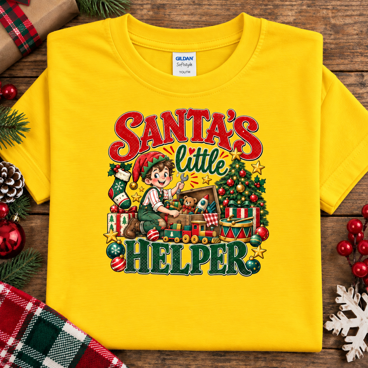 A daisy yellow Gildan Comfort 12x12 short-sleeve t-shirt with a colorful Christmas graphic reading 'Santa's little Helper'. The shirt is displayed flat on a rustic wooden background decorated with pine needles, a pinecone, red berries, and a plaid fabric.