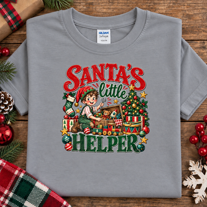 A sport gray Gildan Comfort 100% cotton t-shirt with a colorful graphic print reading 'Santa's Little Helper' featuring an illustration of a child elf working at a toy store. The shirt is displayed flat on a rustic wooden background decorated with Christmas ornaments.