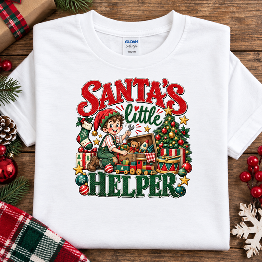 A white Gildan Softstyle youth t-shirt with a colorful Christmas graphic that reads 'Santa's Little Helper'. The shirt is displayed in a flat-lay arrangement on a rustic wooden background, surrounded by holiday decorations.