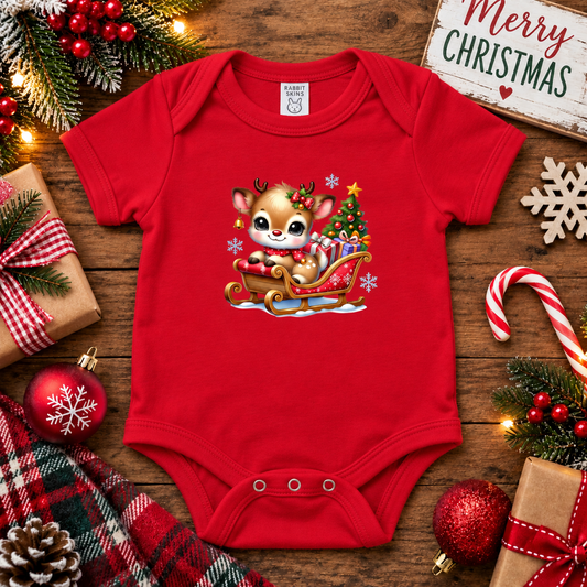 A red short-sleeve baby bodysuit with a colorful Christmas graphic of a cartoon deer in a sleigh by a tree, styled on a wooden surface with holiday decorations including ornaments, candy canes, pine branches, wrapped gifts, and a sign reading 'Merry Christmas'.