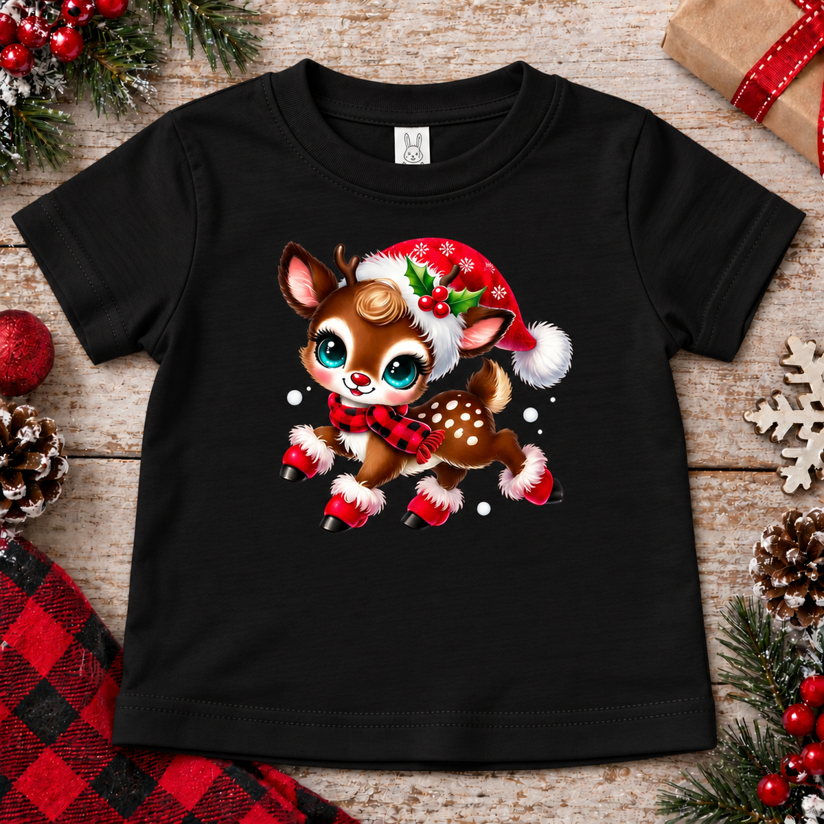 A black toddler short-sleeve t-shirt featuring a cute cartoon Christmas deer graphic, styled in a flat-lay on a rustic wooden background with holiday decorations.
