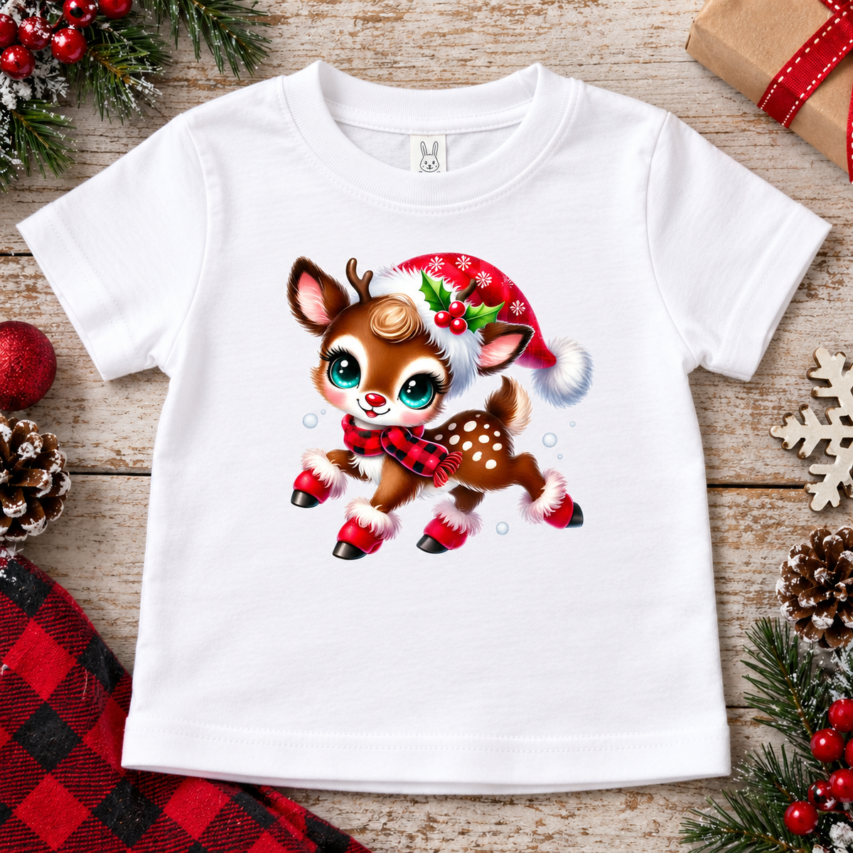 A white short-sleeve baby t-shirt featuring a cute cartoon Christmas deer graphic, styled in a flat-lay on a rustic wooden background with holiday decorations.