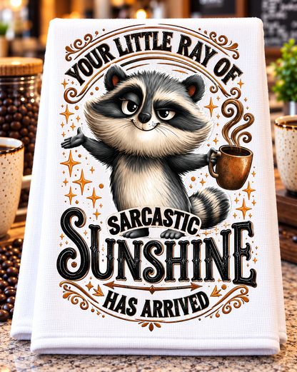 A white kitchen towel with a detailed graphic of a raccoon holding a coffee mug, surrounded by decorative text that reads 'YOUR LITTLE RAY OF SARCASTIC SUNSHINE HAS ARRIVED'. 