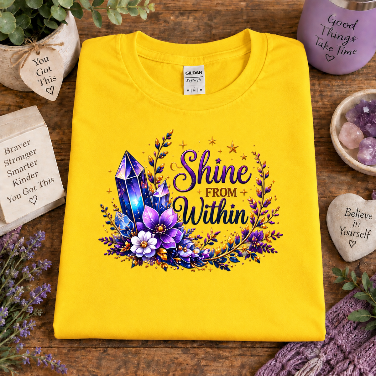 A folded daisy yellow Gildan t-shirt with a colorful graphic reading 'Shine FROM Within' surrounded by purple crystals, flowers, and greenery, styled on a wooden surface with decorative props.