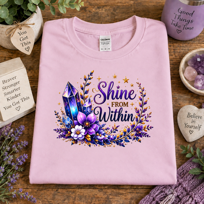 A folded light pink t-shirt with a colorful graphic print reading 'Shine FROM Within' surrounded by purple crystals, flowers, and gold stars, displayed on a wooden table with decorative props.