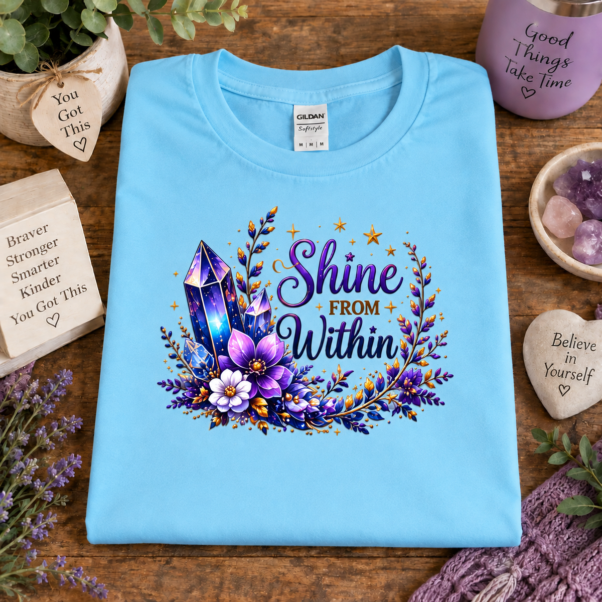 A sky blue folded Gildan t-shirt with a colorful graphic reading 'Shine FROM Within' surrounded by purple crystals, flowers, and gold stars, displayed on a wooden table with decorative props.