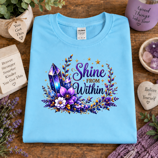 A sky blue folded Gildan t-shirt with a colorful graphic reading 'Shine FROM Within' surrounded by purple crystals, flowers, and gold stars, displayed on a wooden table with decorative props.