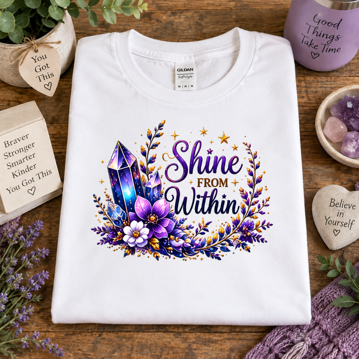 A white Gildan t-shirt is displayed in a flat-lay setting on a wooden table, featuring a colorful graphic with purple crystals, flowers, and the text 'Shine FROM Within'. The shirt is surrounded by decorative props including a purple mug, crystals, and wooden heart tags.