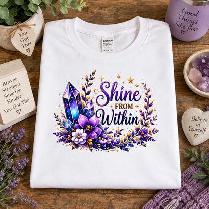 A white Gildan t-shirt is displayed in a flat-lay setting on a wooden table, featuring a colorful graphic with purple crystals, flowers, and the text 'Shine FROM Within'. The shirt is surrounded by decorative props including a purple mug, crystals, and wooden heart tags.