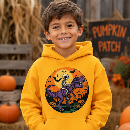 A smiling young boy wearing a bright yellow hoodie with a circular Halloween graphic of a skeleton riding a purple dinosaur. He is posed outdoors in a fall-themed setting with pumpkins, hay bales, and a 'Pumpkin Patch' sign in the background.