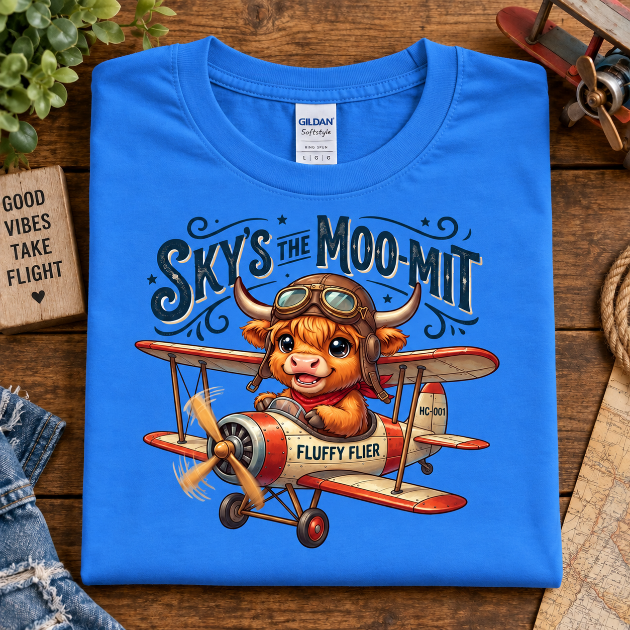 A folded heather royal blue Gildan Softstyle T-shirt is displayed on a rustic wooden surface. The front of the shirt features a colorful graphic of a cartoon Scottish Highland cow wearing aviator goggles and driving a vintage red and cream biplane, with the text 'SKY'S THE MOO-MIT' and 'FLUFFY FLIER'.