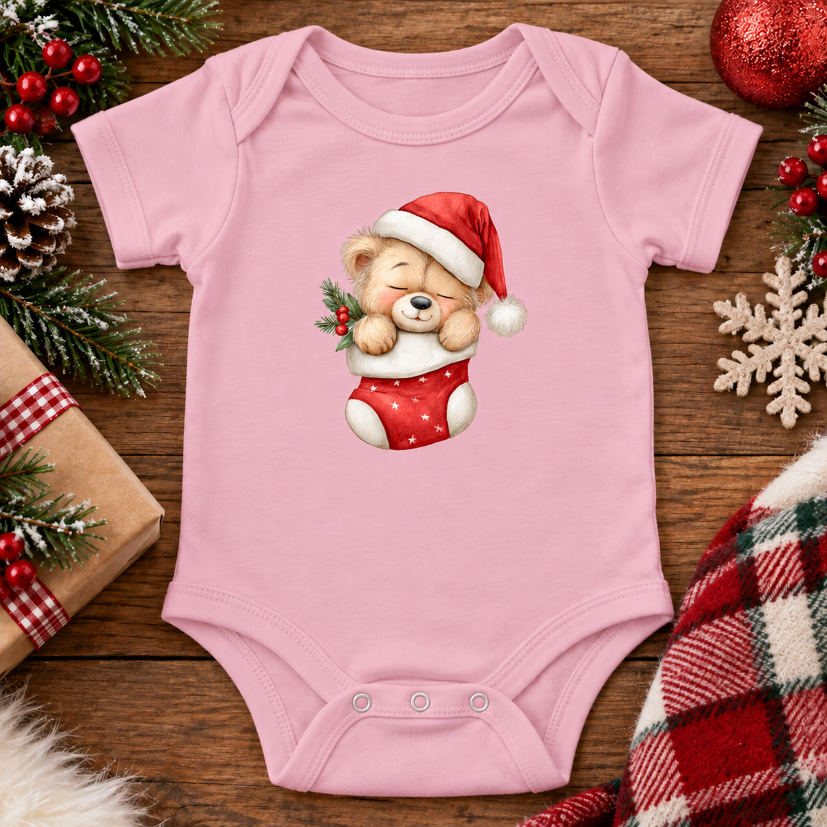A light pink baby bodysuit with a cute teddy bear graphic wearing a Santa hat and red stocking, displayed on a rustic wooden background with Christmas decorations.