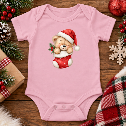 A light pink baby bodysuit with a cute teddy bear graphic wearing a Santa hat and red stocking, displayed on a rustic wooden background with Christmas decorations.