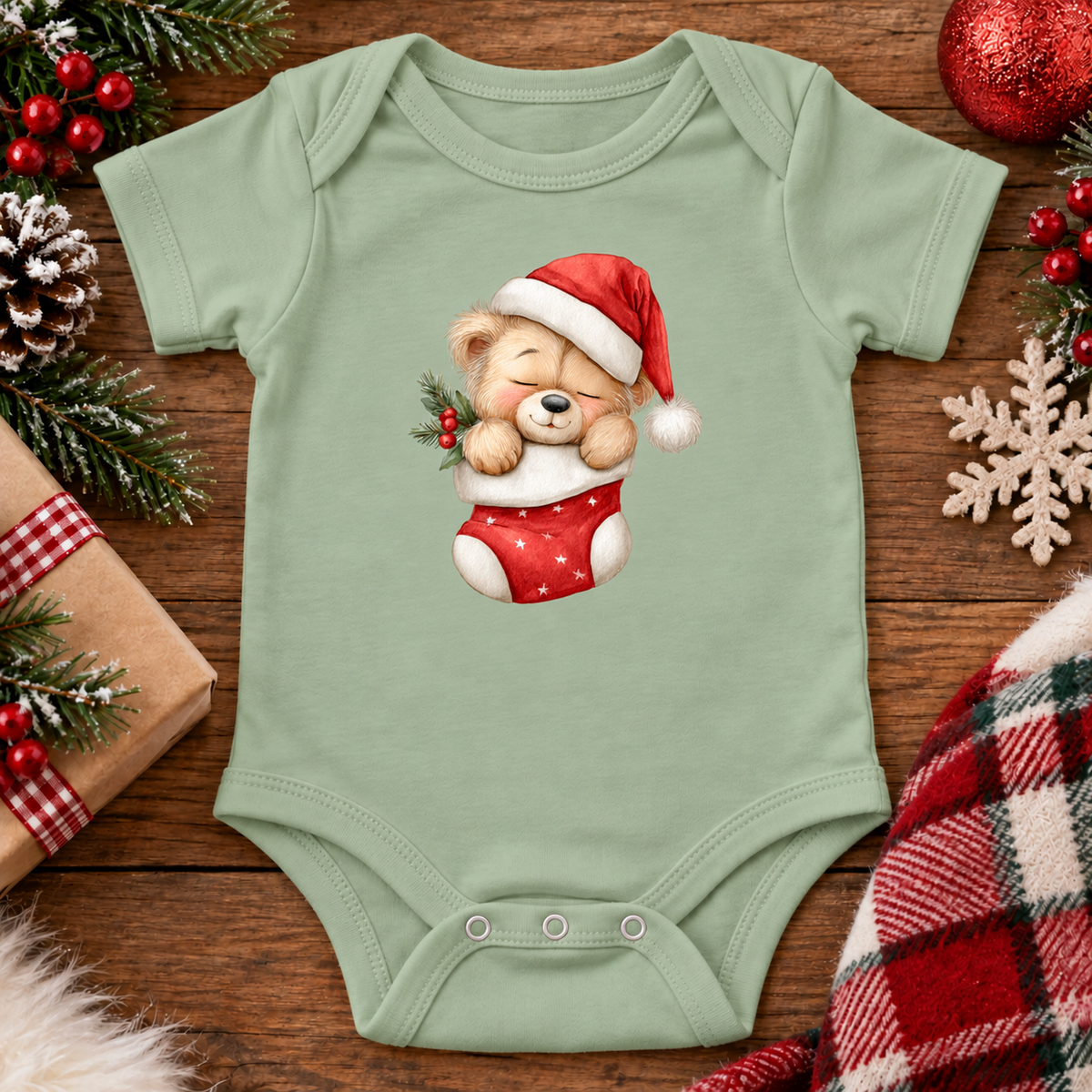A sage green baby bodysuit with a festive Christmas graphic of a teddy bear in a Santa hat and stocking, displayed on a wooden surface with holiday decorations.