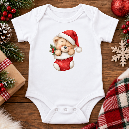 A white short-sleeve baby bodysuit laid flat on a wooden surface, featuring a printed illustration of a teddy bear wearing a red Santa hat and red stocking. The bodysuit is styled with Christmas-themed props including pine branches, red berries, a wrapped gift, a plaid fabric, and a snowflake ornament.