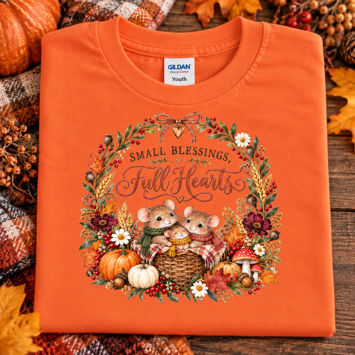 An orange Gildan Youth t-shirt is laid flat on a rustic wooden surface surrounded by autumn decorations. The front of the shirt features a detailed graphic of two mice in a basket surrounded by pumpkins, fall leaves, and the text 'SMALL BLESSINGS, Full Hearts'.