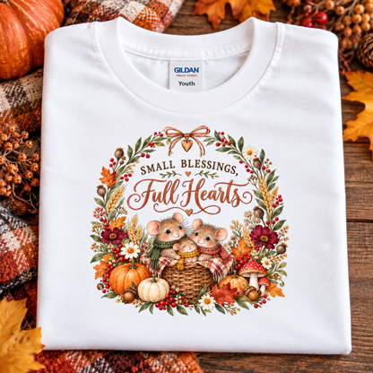 A folded white Gildan Youth t-shirt with a 'Small Blessings, Full Hearts' autumn mouse graphic, displayed on a rustic wooden surface surrounded by fall leaves, pumpkins, and a plaid blanket.