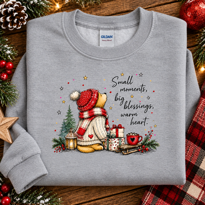 A folded sport gray Gildan Heavy Blend sweatshirt with a festive teddy bear Christmas graphic and the text 'Small moments, big blessings, warm heart.' surrounded by holiday decorations on a wooden surface.