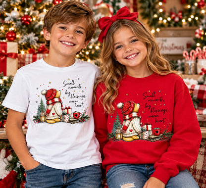 A young boy and girl wearing matching Christmas-themed graphic t-shirt and sweatshirt sets, posed together in front of a decorated Christmas tree and holiday ornaments. The boy wears a white t-shirt and the girl wears a red sweatshirt, both featuring a cartoon snowman graphic and the text 'Small moments, big blessings, warm heart'.