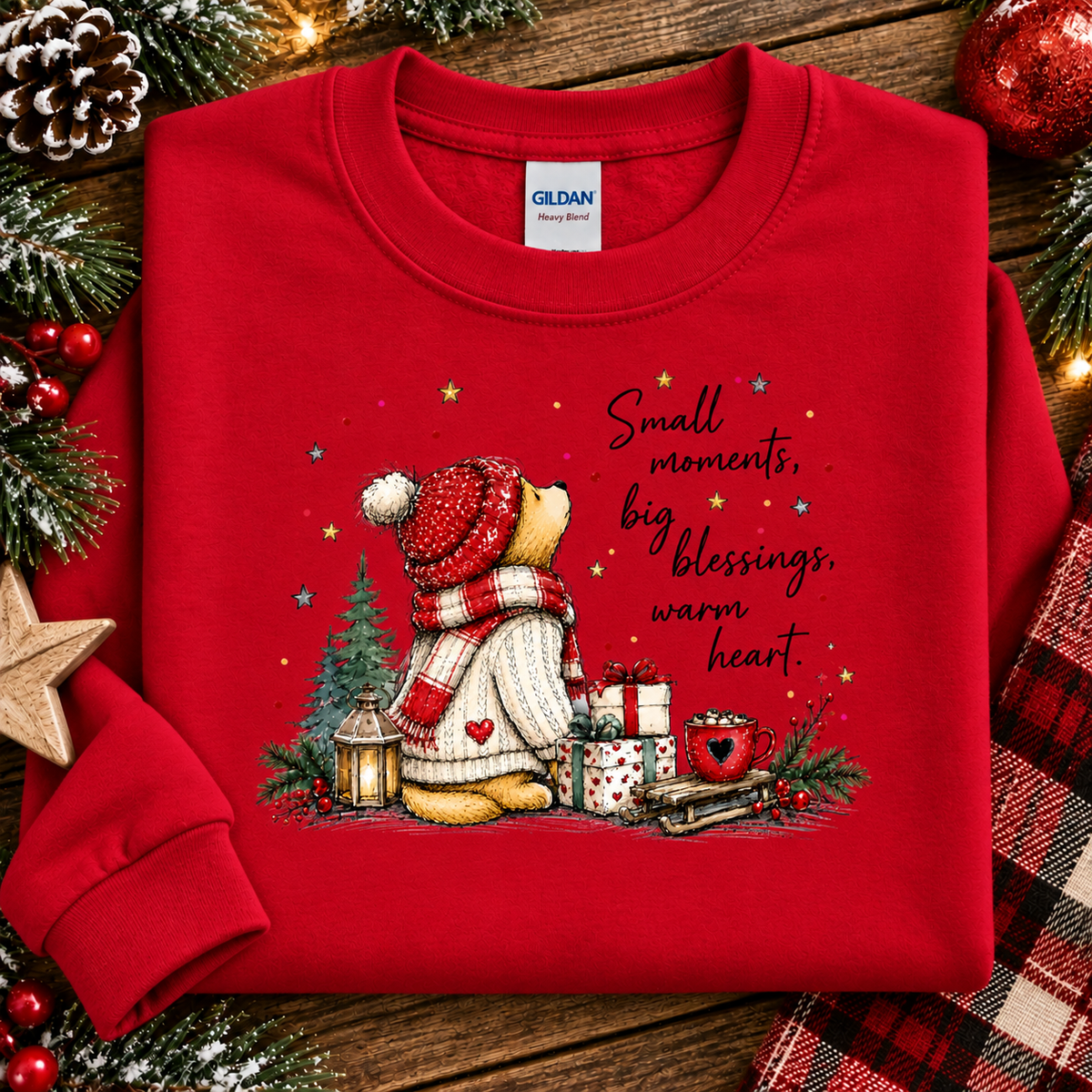 A folded red crewneck sweatshirt by Gildan, featuring a festive Christmas graphic of a teddy bear in a red hat and scarf with the text 'Small moments, big blessings, warm heart.' The sweatshirt is styled on a rustic wooden background with holiday decorations including pine branches, a star, and a plaid scarf.