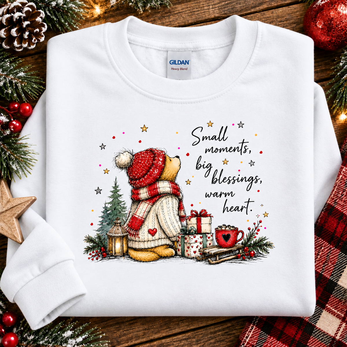 A folded white Gildan crewneck sweatshirt with a festive Christmas graphic, displayed on a rustic wooden background decorated with pine branches, a pinecone, red berries, and a red plaid cloth. The graphic features a teddy bear in a red knit hat and scarf next to a mug with the text 'Small moments, big blessings, warm heart.