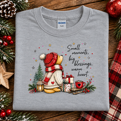 A folded sport gray Gildan Youth t-shirt with a Christmas-themed graphic of a teddy bear and a holiday quote, displayed on a rustic wooden background with pine branches and red berries.