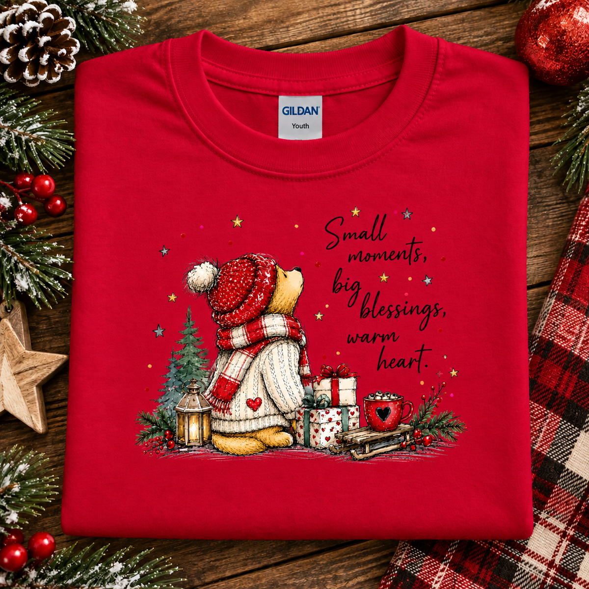 A folded red Gildan Youth t-shirt with a Christmas-themed graphic of a teddy bear wearing a red and white striped beanie and scarf, sitting by a Christmas tree with gifts and the text 'Small moments, big blessings, warm heart.' The shirt is styled on a rustic wooden background with pine branches, a pinecone, a star, and a plaid fabric.