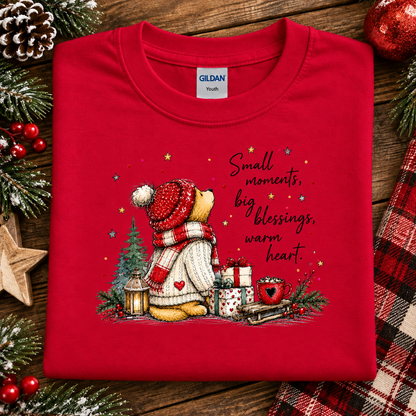 A folded red Gildan Youth t-shirt with a Christmas-themed graphic of a teddy bear wearing a red and white striped beanie and scarf, sitting by a Christmas tree with gifts and the text 'Small moments, big blessings, warm heart.' The shirt is styled on a rustic wooden background with pine branches, a pinecone, a star, and a plaid fabric.