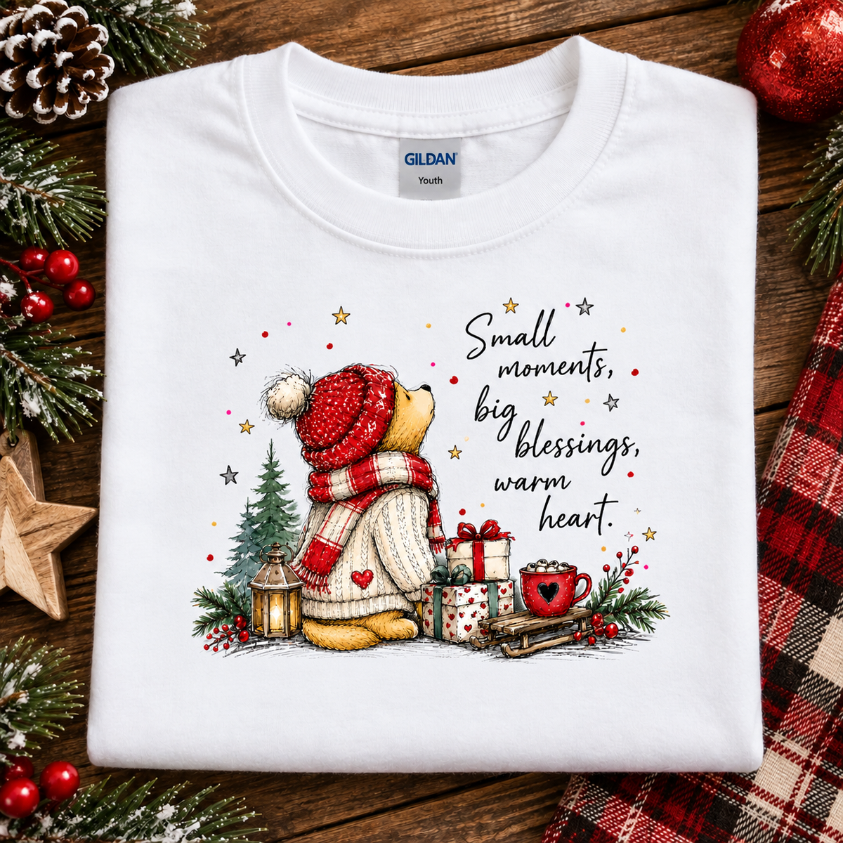 A folded white Gildan Youth t-shirt with a festive Christmas graphic, displayed on a rustic wooden background with holiday decorations like pine branches, red berries, and a plaid scarf.