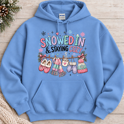 A carolina blue colored hoodie with a festive winter graphic with the phrase “SNOWED IN & STAYING COZY” in pastel lettering, surrounded by snowflakes and decorated with cozy items including a scarf, mittens, slippers, a mug of hot cocoa, and hats hanging from a clothesline.