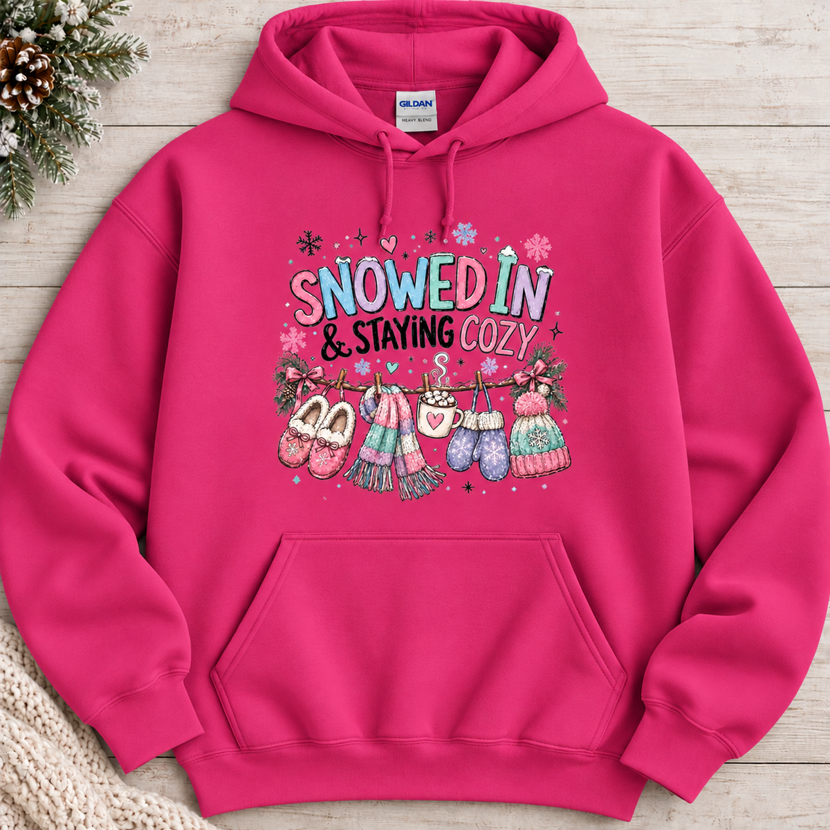 A heliconia pink hoodie with a festive winter graphic with the phrase “SNOWED IN & STAYING COZY” in pastel lettering, surrounded by snowflakes and decorated with cozy items including a scarf, mittens, slippers, a mug of hot cocoa, and hats hanging from a clothesline.