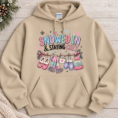 A sand colored hoodie with a festive winter graphic with the phrase “SNOWED IN & STAYING COZY” in pastel lettering, surrounded by snowflakes and decorated with cozy items including a scarf, mittens, slippers, a mug of hot cocoa, and hats hanging from a clothesline.
