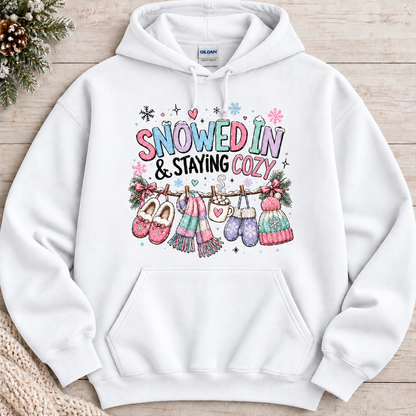 A white hoodie with a festive winter graphic with the phrase “SNOWED IN & STAYING COZY” in pastel lettering, surrounded by snowflakes and decorated with cozy items including a scarf, mittens, slippers, a mug of hot cocoa, and hats hanging from a clothesline.