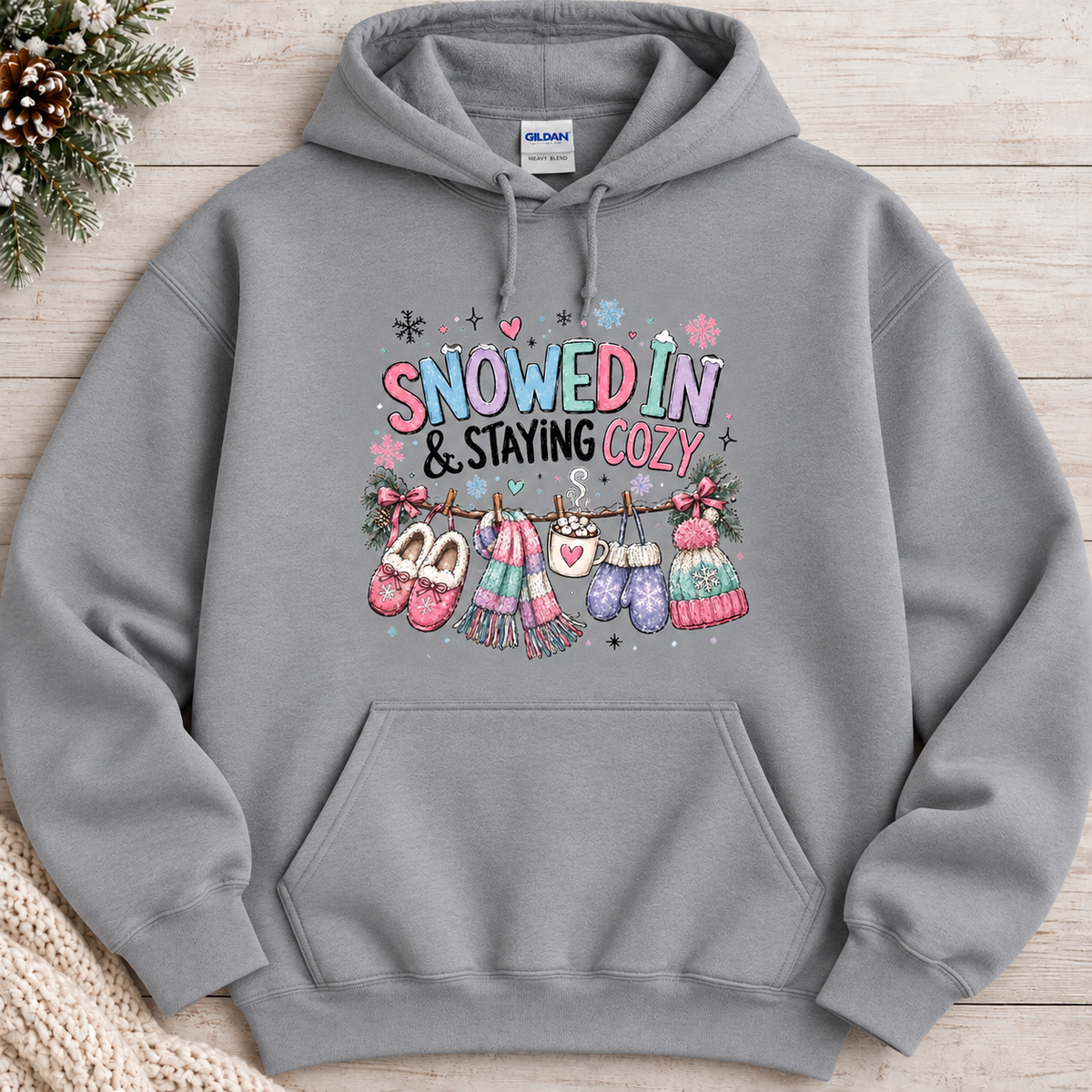A sport grey colored hoodie with a festive winter graphic with the phrase “SNOWED IN & STAYING COZY” in pastel lettering, surrounded by snowflakes and decorated with cozy items including a scarf, mittens, slippers, a mug of hot cocoa, and hats hanging from a clothesline.