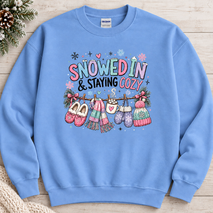 A carolina blue colored sweatshirt with a festive winter graphic with the phrase “SNOWED IN & STAYING COZY” in pastel lettering, surrounded by snowflakes and decorated with cozy items including a scarf, mittens, slippers, a mug of hot cocoa, and hats hanging from a clothesline.