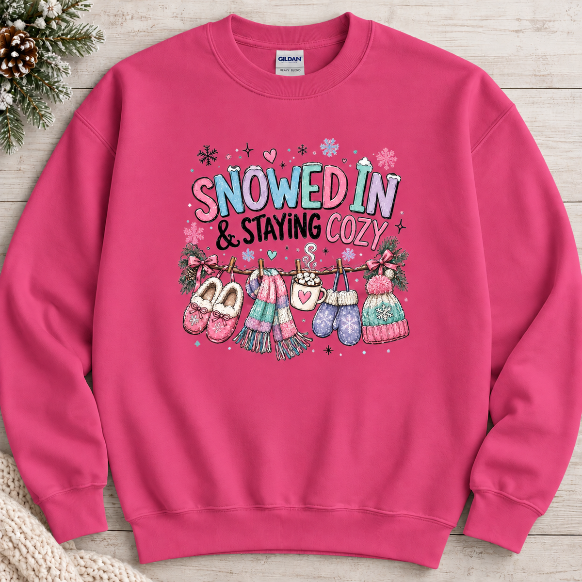 A heliconia pink colored sweatshirt with a festive winter graphic with the phrase “SNOWED IN & STAYING COZY” in pastel lettering, surrounded by snowflakes and decorated with cozy items including a scarf, mittens, slippers, a mug of hot cocoa, and hats hanging from a clothesline.