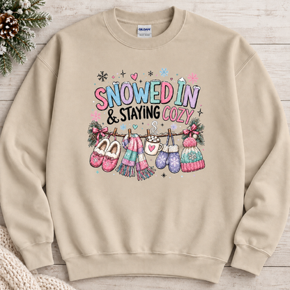 A sand colored sweatshirt with a festive winter graphic with the phrase “SNOWED IN & STAYING COZY” in pastel lettering, surrounded by snowflakes and decorated with cozy items including a scarf, mittens, slippers, a mug of hot cocoa, and hats hanging from a clothesline.