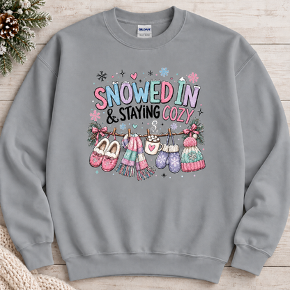 A sport grey colored sweatshirt with a festive winter graphic with the phrase “SNOWED IN & STAYING COZY” in pastel lettering, surrounded by snowflakes and decorated with cozy items including a scarf, mittens, slippers, a mug of hot cocoa, and hats hanging from a clothesline.