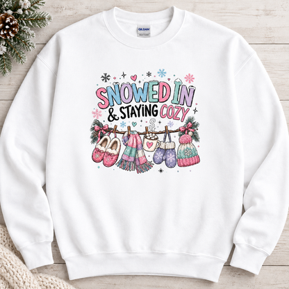 A white sweatshirt with a festive winter graphic with the phrase “SNOWED IN & STAYING COZY” in pastel lettering, surrounded by snowflakes and decorated with cozy items including a scarf, mittens, slippers, a mug of hot cocoa, and hats hanging from a clothesline.