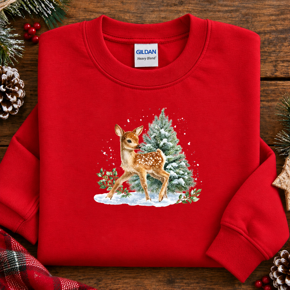 A folded red crewneck sweatshirt with a Christmas graphic of a deer and a snowy tree, styled on a wooden background with pine branches and red berries. The Gildan brand label is visible inside the collar.