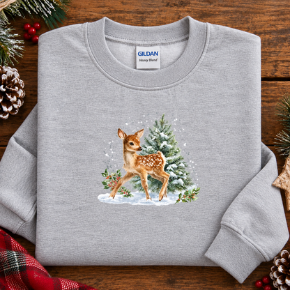 A folded sport gray Gildan sweatshirt on a rustic wooden surface, decorated with pine branches, red berries, and a red plaid cloth. The sweatshirt features a graphic of a brown fawn standing next to a snowy green pine tree.